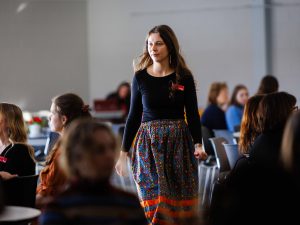 A young woman wearing a name tag and patterned skirt walks between tables as attendees listen during a REDress Day event.