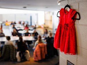A small red dress hangs on a wall in the foreground, symbolizing Missing and Murdered Indigenous Women and Girls, while participants gather in the background.