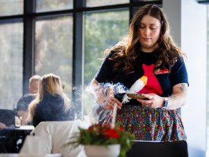 An organizer holds a smoldering bundle of sage and a feather, performing a smudging ceremony inside a sunlit room.