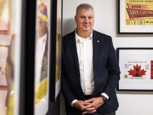 A man poses for a photo standing against a wall beside several framed images.