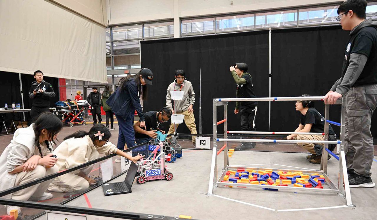 A group of students work on computers and with robots while preparing them to navigate a competition space with colourful blocks.