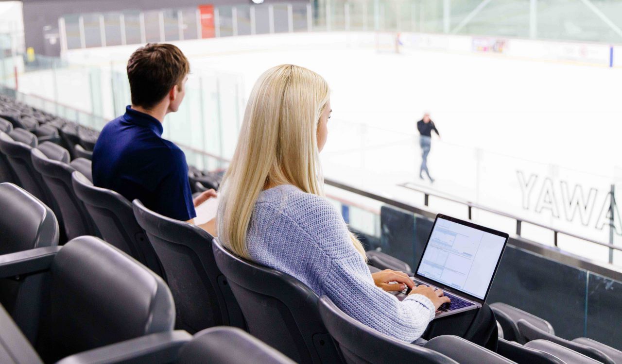 Two people work on laptops while observing activity in an ice rink.