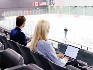 Two people work on laptops while observing activity in an ice rink.