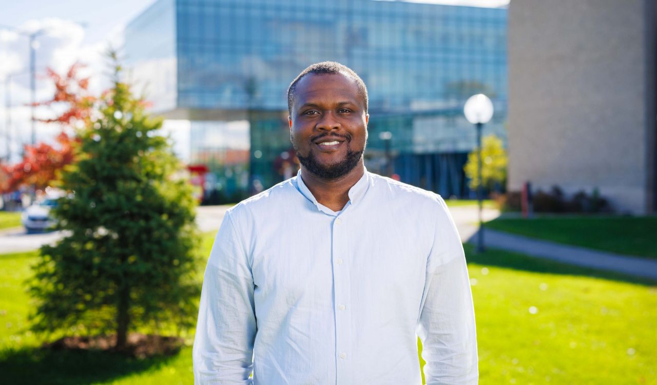 A man stands outside on a university campus with green grass, trees and campus buildings in the background.