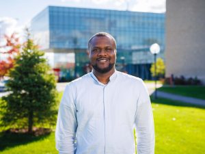 A man stands outside on a university campus with green grass, trees and campus buildings in the background.