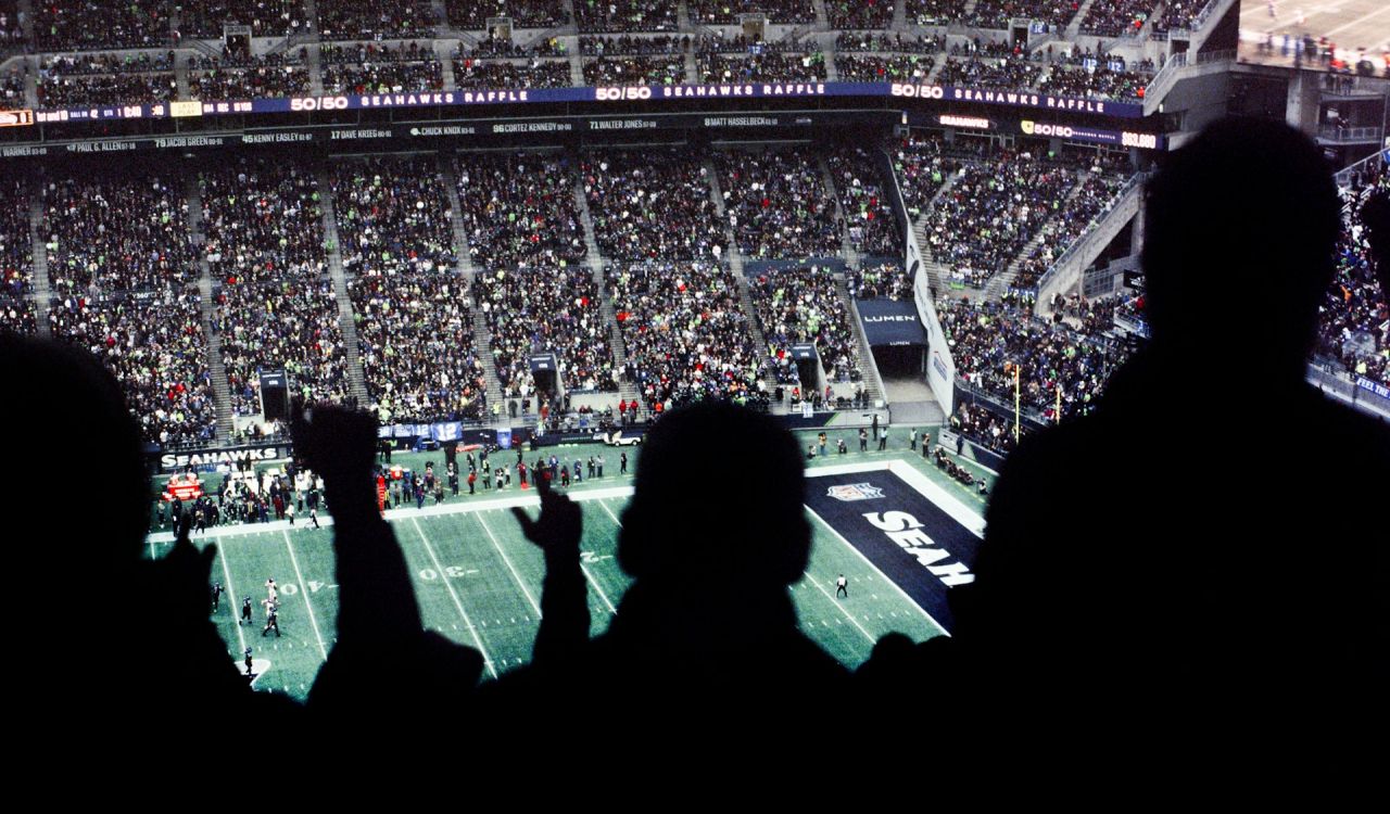 A crowd cheers at a football game with the field and stadium in the background.