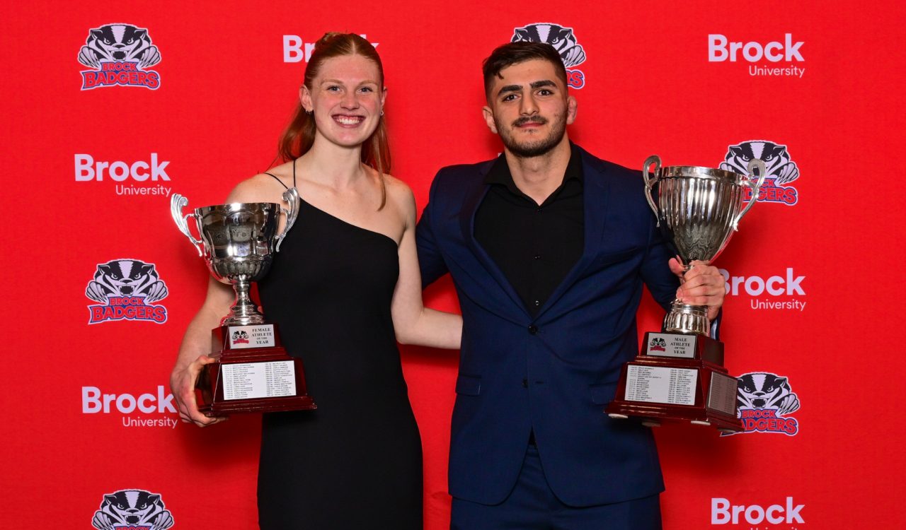 Two university students in formal wear pose with trophies against a red backdrop.