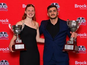 Two university students in formal wear pose with trophies against a red backdrop.
