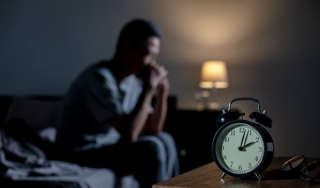 Tired sits on bed in dark room with alarm clock in the foreground.