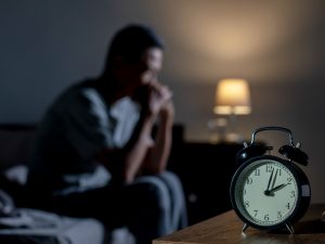 Tired sits on bed in dark room with alarm clock in the foreground.