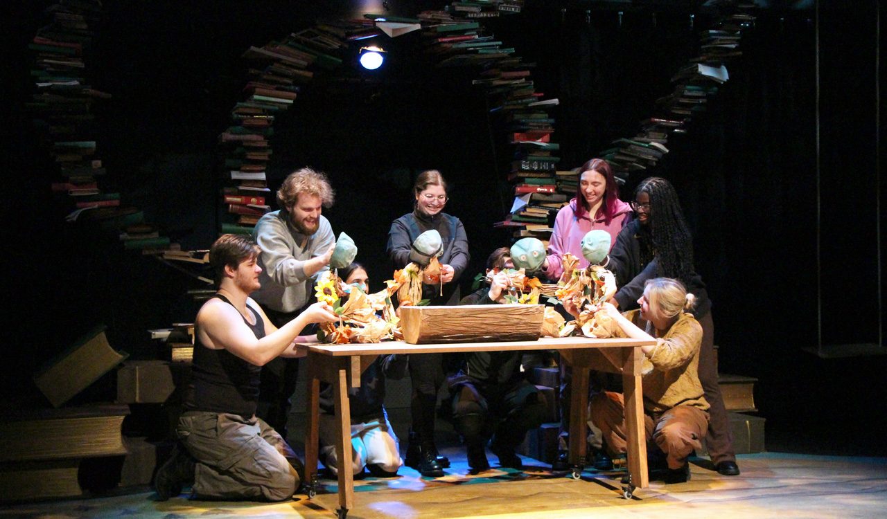Group of people sit around a wooden table with stacks of books behind them.