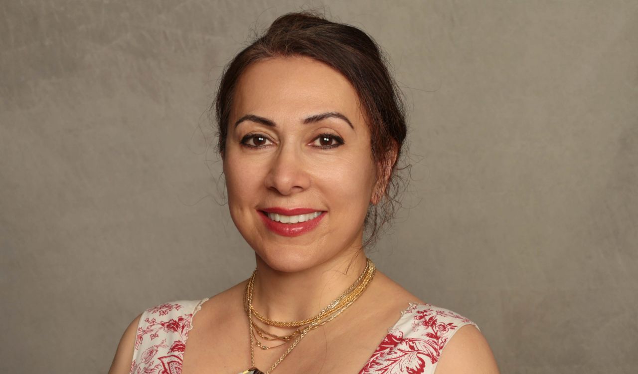 A woman smiles at the camera while sitting in front of a tan backdrop.