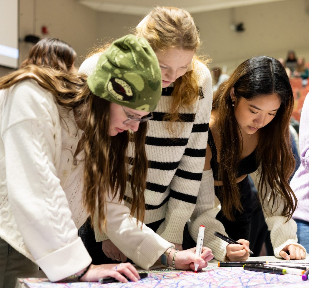 Three students use markers to colour on white paper across a table.