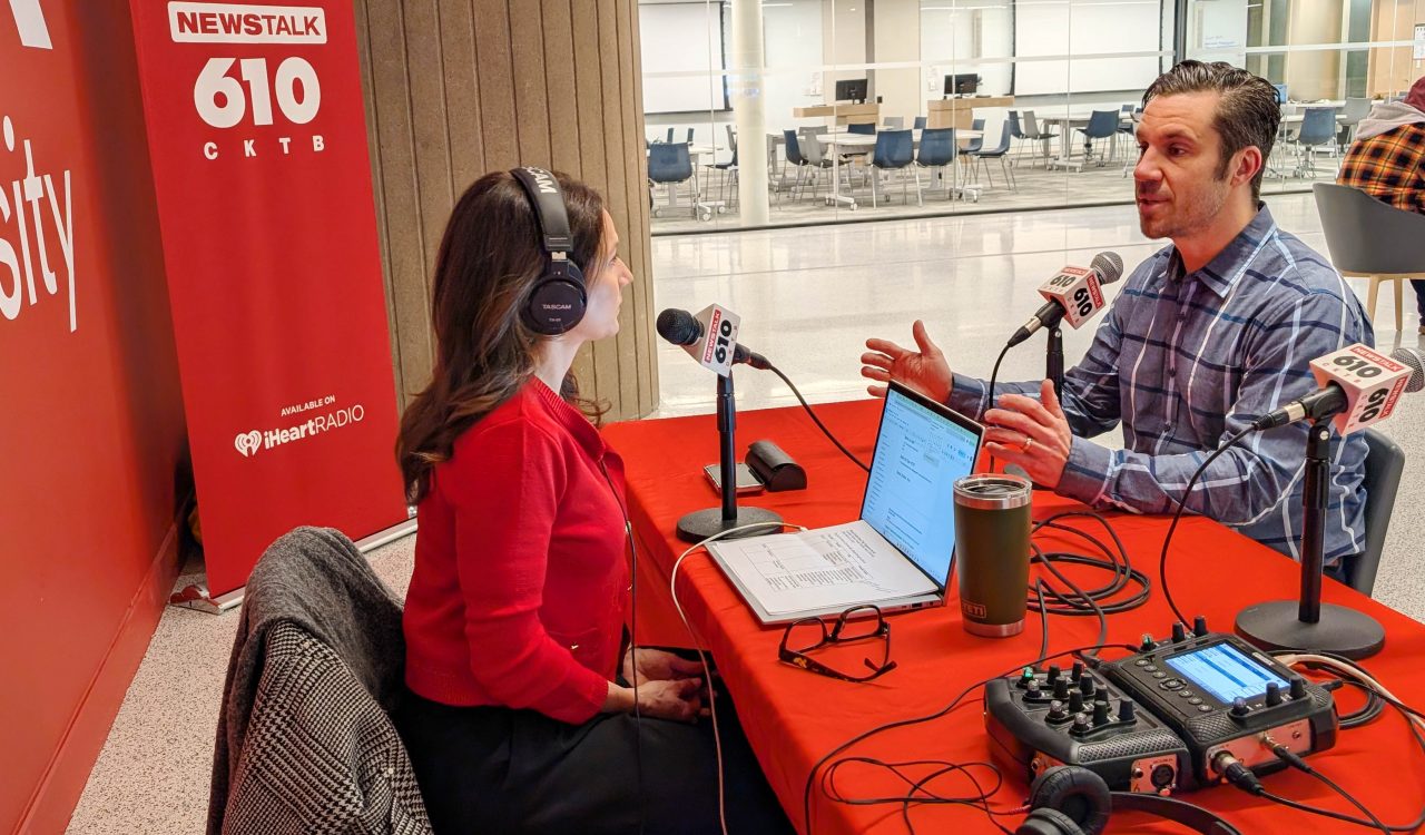 Two people conduct a radio interview, speaking into microphones while seated at a table.