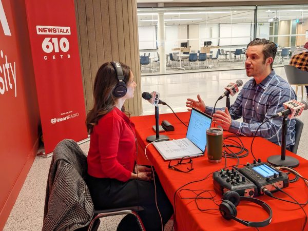 Two people conduct a radio interview, speaking into microphones while seated at a table.