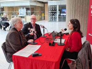 Three people conduct a radio interview while sitting at a table and speaking into microphones.