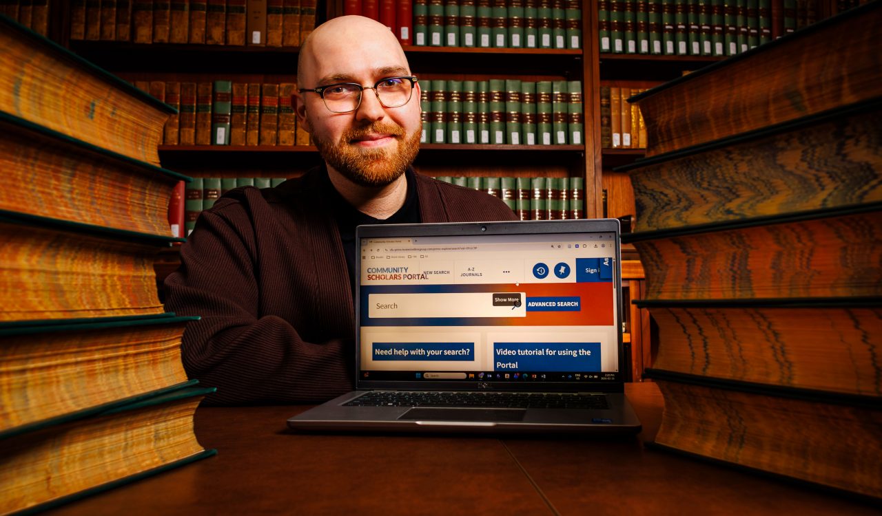 Man sits at a desk behind a laptop and positioned between stacks of books.
