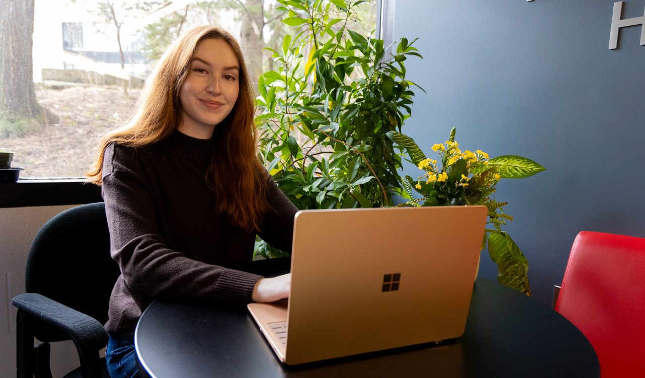 A young woman works on a laptop in front of a window.