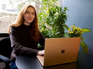 A young woman works on a laptop in front of a window.