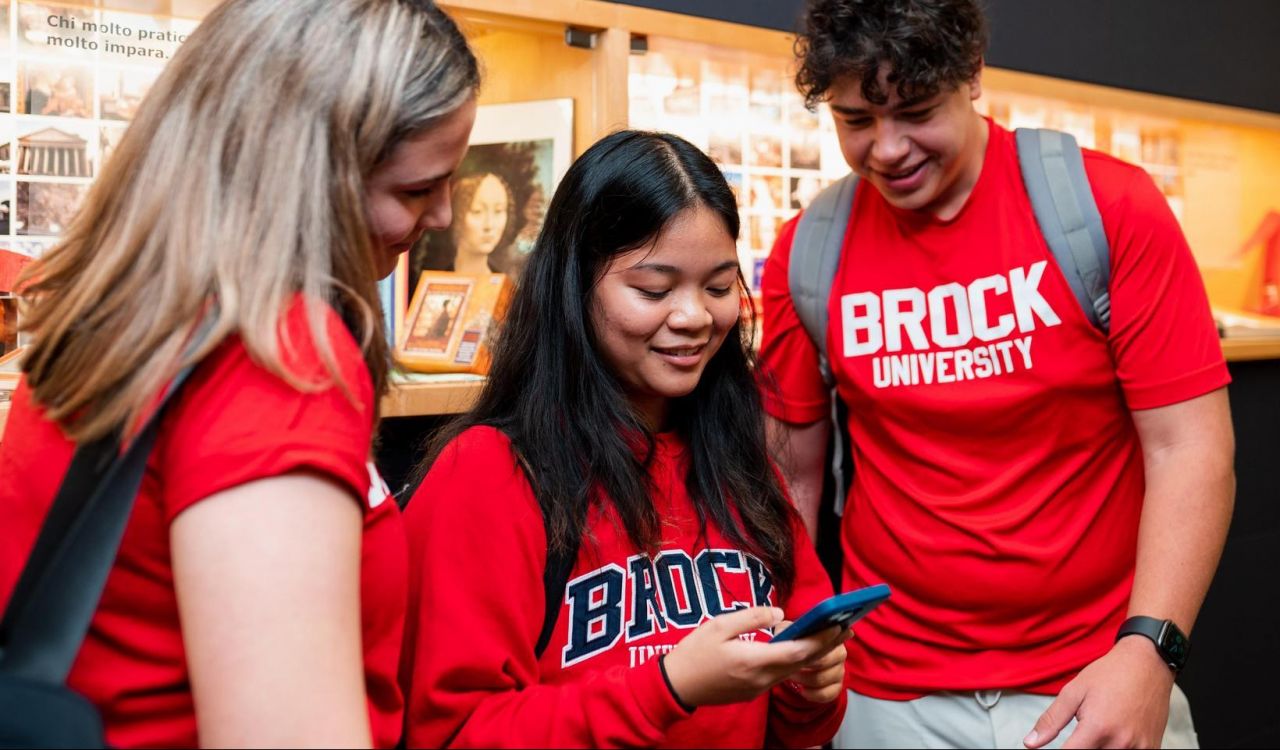 Three students wearing red Brock University shirts stand together indoors, looking at a mobile phone one of them is holding. A display case with memorabilia is visible in the background.