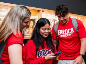 Three students wearing red Brock University shirts stand together indoors, looking at a mobile phone one of them is holding. A display case with memorabilia is visible in the background.