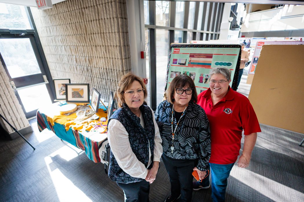 Three people stand together smiling beside a table of Indigenous ribbon skirt materials.
