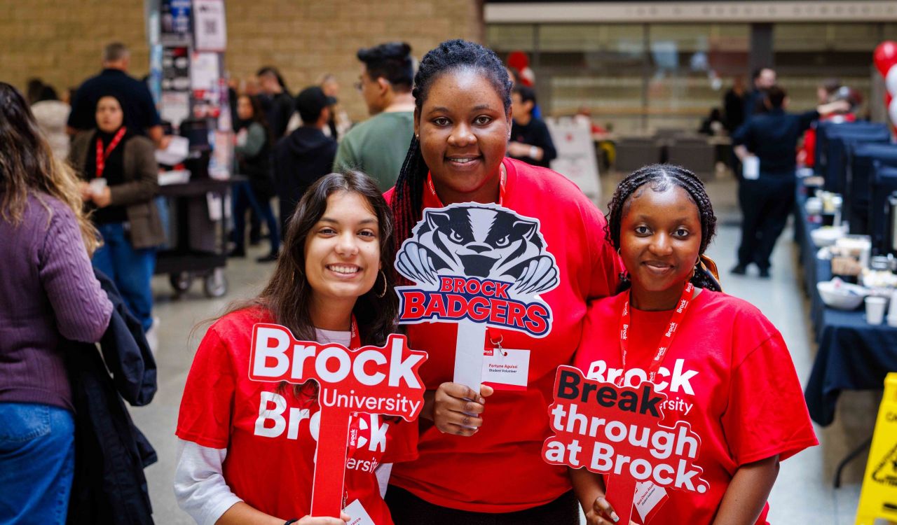 Three students in Brock University gear pose with photo prop signs at an information fair.