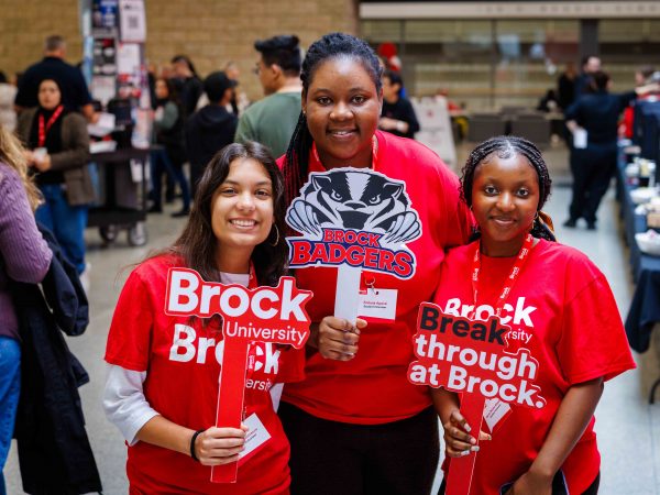 Three students in Brock University gear pose with photo prop signs at an information fair.
