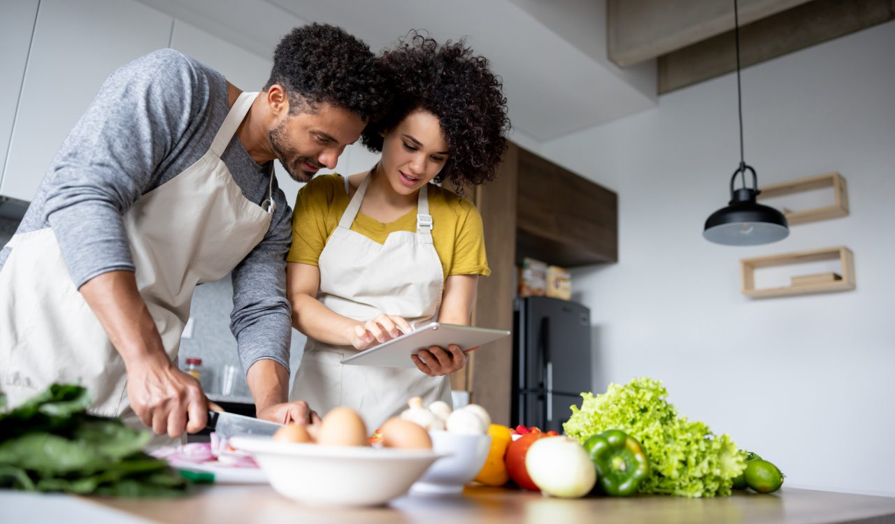 Two young people look at a tablet in a kitchen while cooking vegetables.
