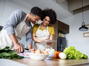 Two young people look at a tablet in a kitchen while cooking vegetables.