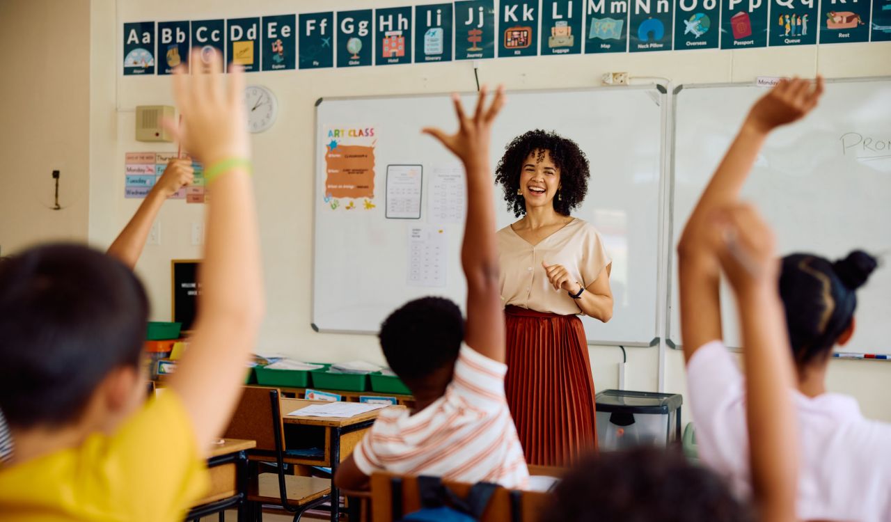 A teacher smiles at a group of students in a classroom who are raising their hands.