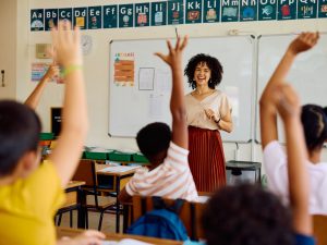 A teacher smiles at a group of students in a classroom who are raising their hands.