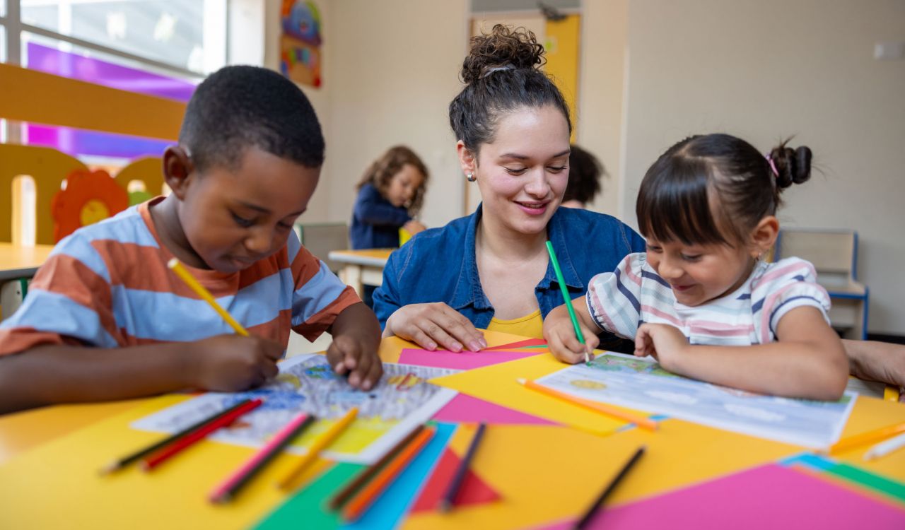 A woman sits with young students doing crafts in a primary classroom.