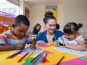 A woman sits with young students doing crafts in a primary classroom.