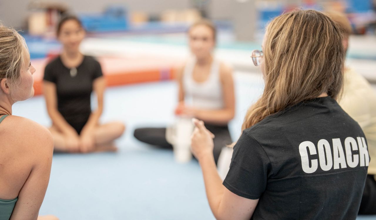 A coach, seen from behind, talks to a group of athletes seated on the floor in a circle.