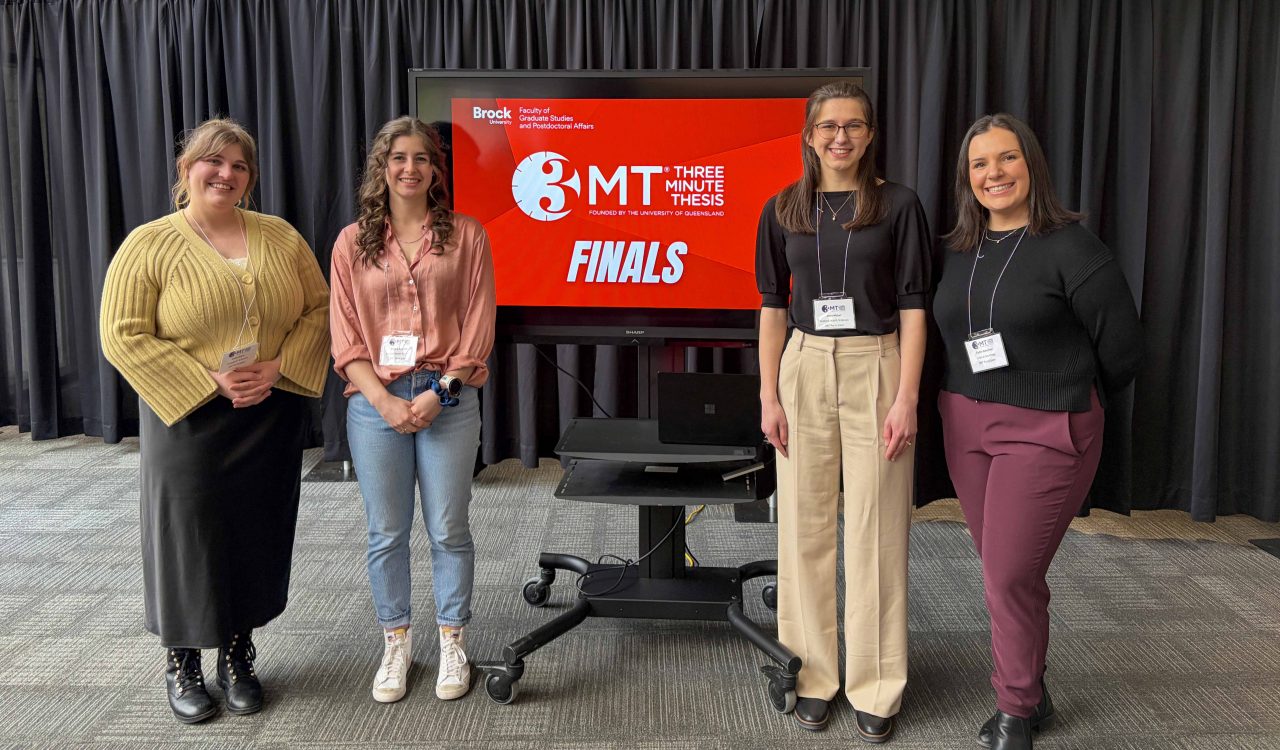 Four university students pose beside a screen displaying the 3MT logo.