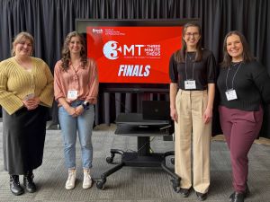 Four university students pose beside a screen displaying the 3MT logo.