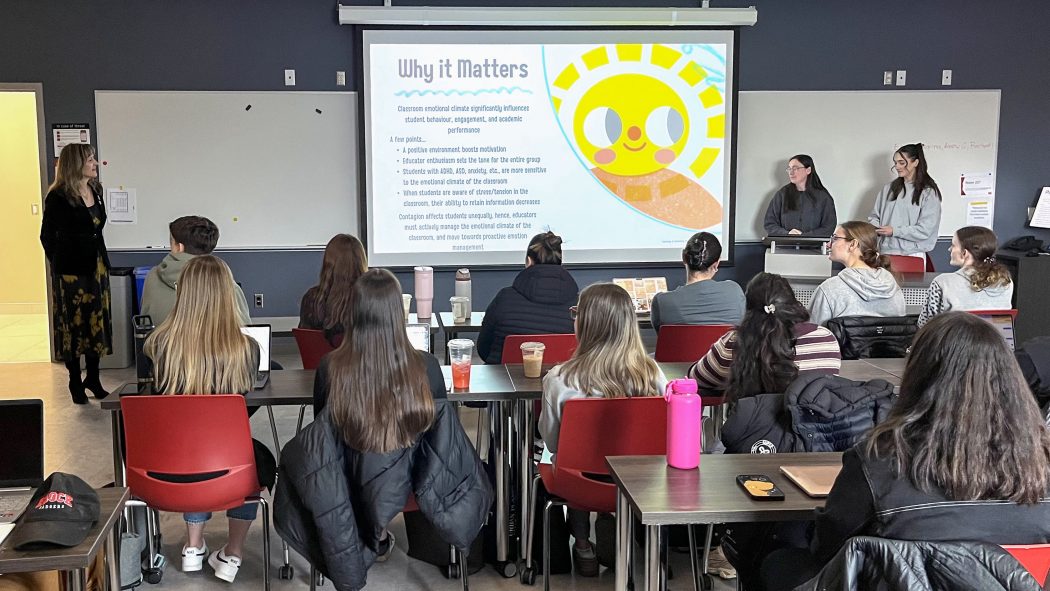 Three people stand by a screen at the front of a university classroom.