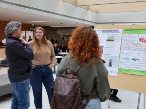 Three people look at a poster in a sunny atrium.