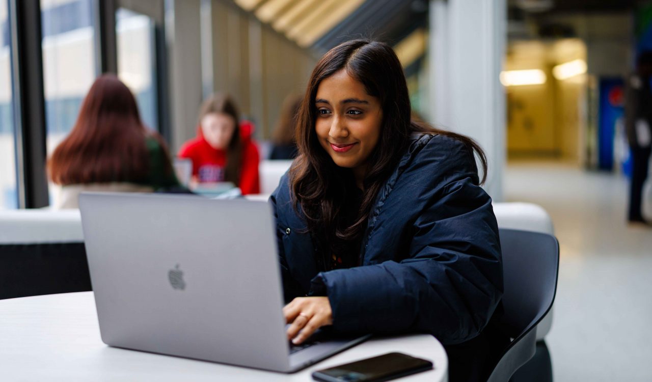 A person sitting at a table indoors, working on a laptop with a smartphone placed nearby. Other people are seated in the background, and large windows line the space.