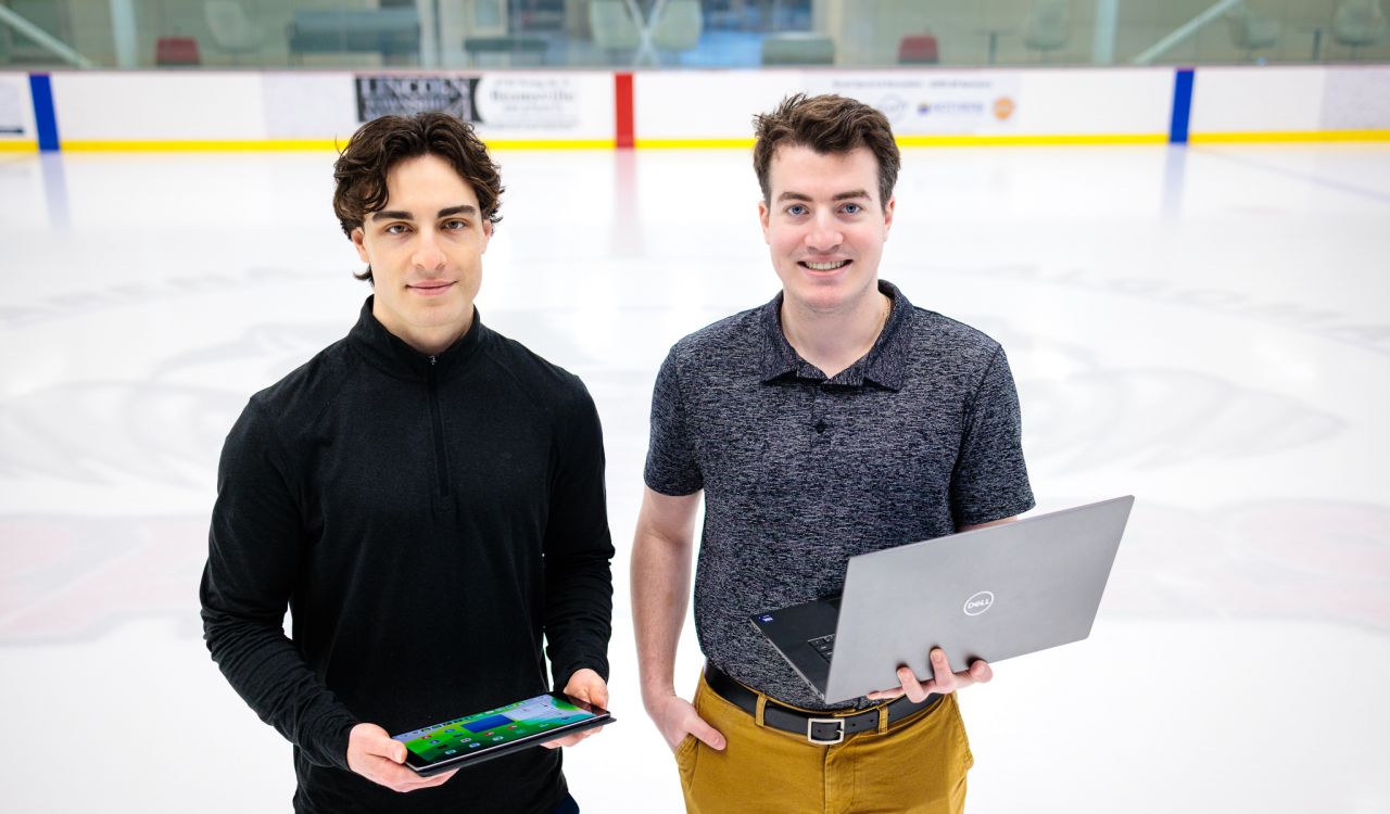 Two young men — the man on the left holding a tablet and the man on the right holding a laptop — stand side-by-side on the ice of a hockey rink with billboards fuzzed out in the background.
