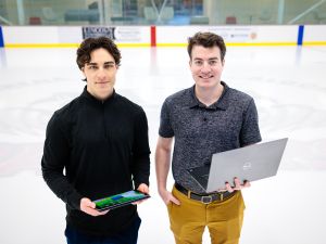 Two young men — the man on the left holding a tablet and the man on the right holding a laptop — stand side-by-side on the ice of a hockey rink with billboards fuzzed out in the background.