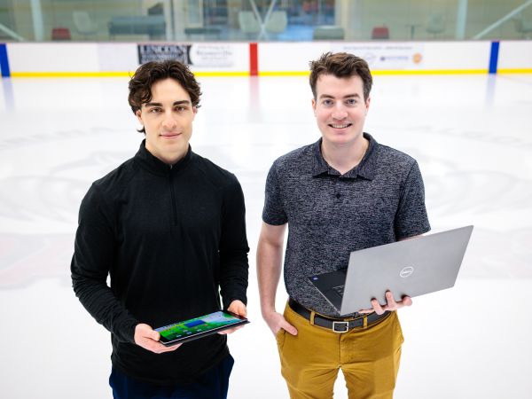 Two young men — the man on the left holding a tablet and the man on the right holding a laptop — stand side-by-side on the ice of a hockey rink with billboards fuzzed out in the background.