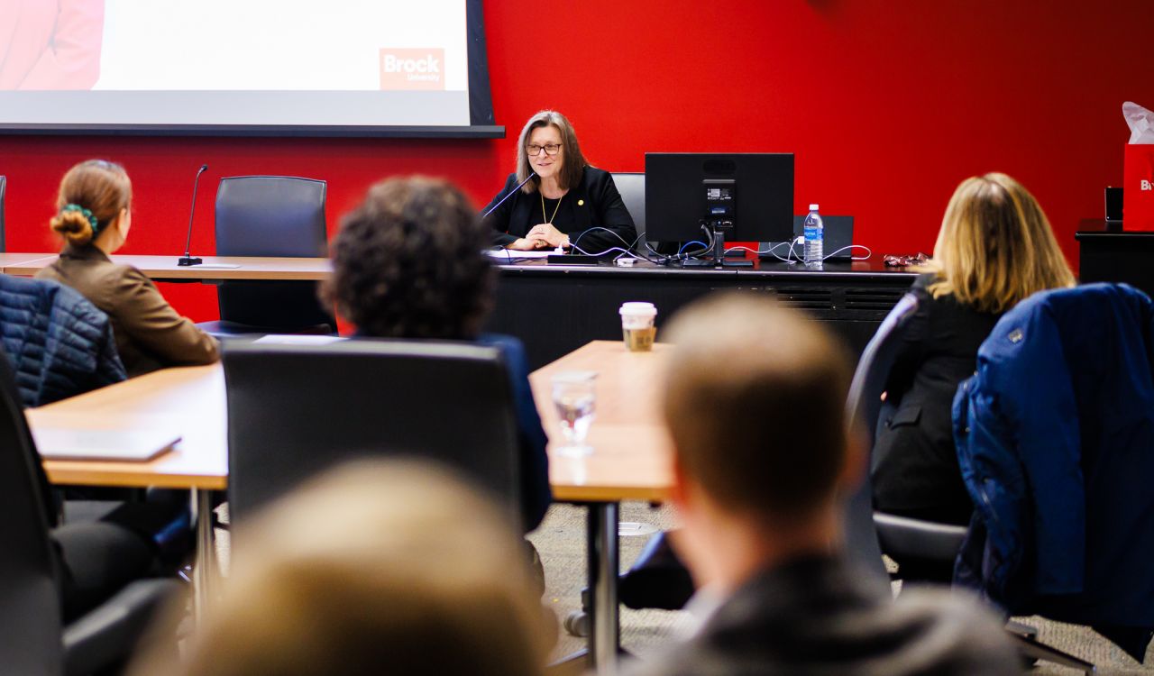 A woman sits at table and speaks into a microphone in front of a group of event attendees.