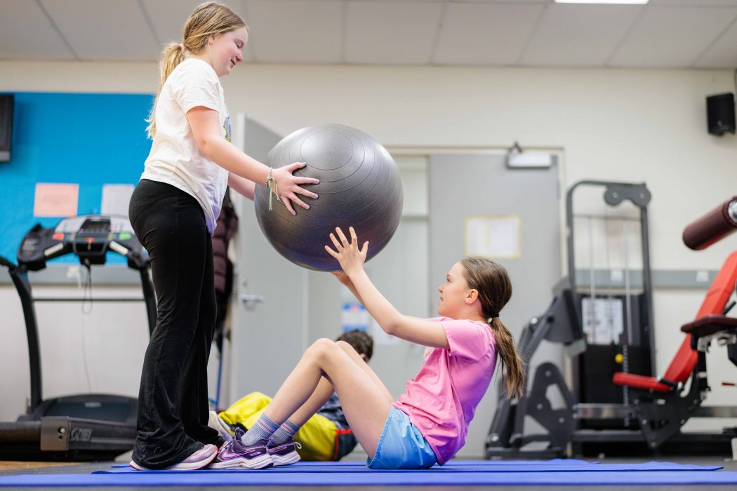 A young woman hands a girl, who is exercising on the ground, a large inflated ball.