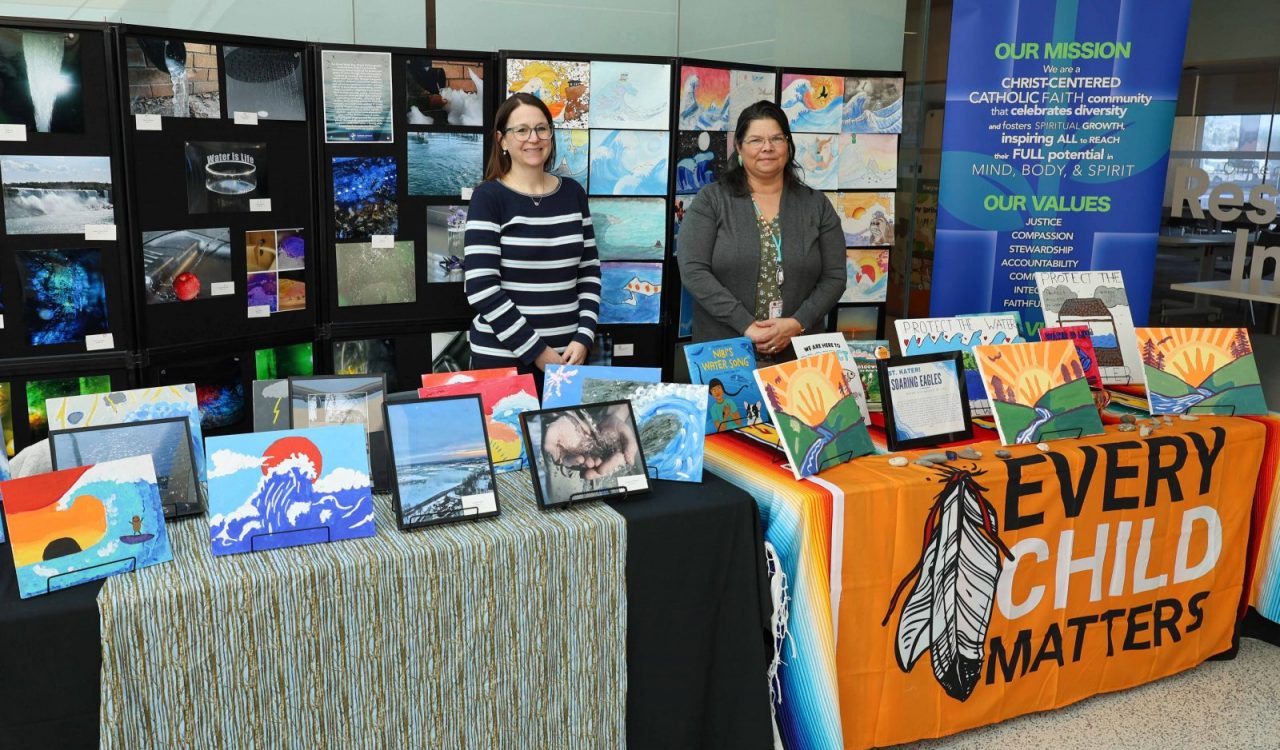 Two women stand behind tables showcasing art created by children and youth.