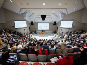 People sitting in a large auditorium listen to a speaker at a university open house event.