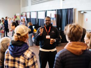 A man speaks to a group of guests at a university information fair.