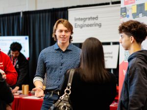 Two people have a conversation in a busy room at a university open house event.