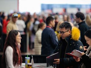 Two people have a conversation in a busy room at a university open house event.